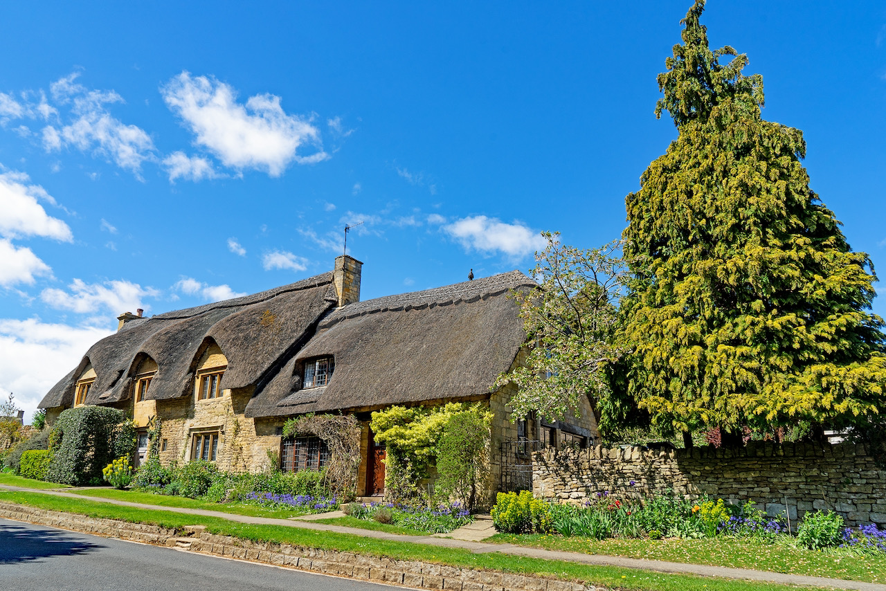 Long Cotswold stone cottage with a thatched roof, eyebrow dormer windows and a stone chimney, behind a dry-stone wall